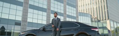 Proud car owner posing at black classic automobile on modern street. Elegant bearded businessman standing near auto premium class outdoor. Rich serious ceo manager crossing hands expressing confidence