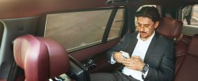 Young elegant businessman sitting on the back seat of a luxury car and using smartphone close up